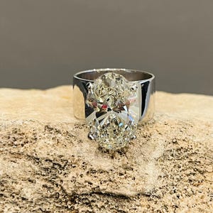 May include: A silver ring with a large oval-shaped diamond set in the center. The ring is on a rough, light brown surface.