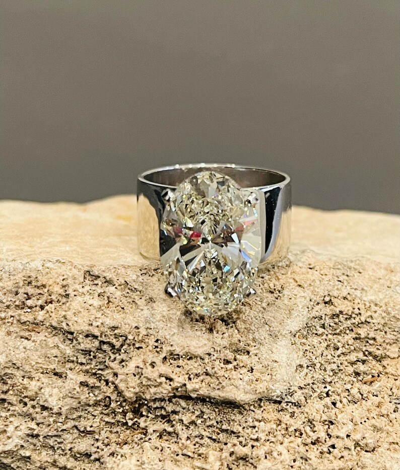 May include: A silver ring with a large oval-shaped diamond set in the center. The ring is on a rough, light brown surface.