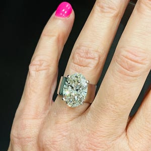 May include: A large oval-shaped diamond engagement ring set in a white gold band. The ring has a wide band and the diamond is surrounded by smaller diamonds.