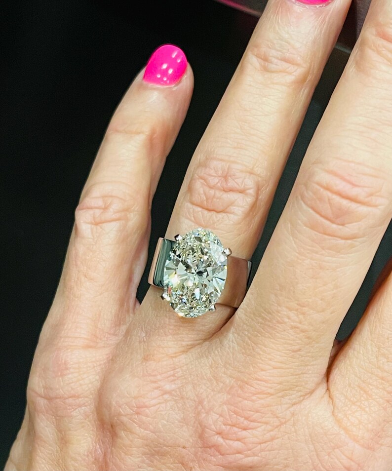 May include: A large oval-shaped diamond engagement ring set in a white gold band. The ring has a wide band and the diamond is surrounded by smaller diamonds.