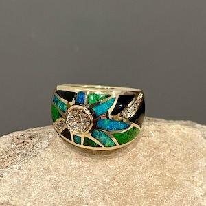 May include: A gold ring with a black, green, and blue inlay design. The ring features a round, clear stone in the center.