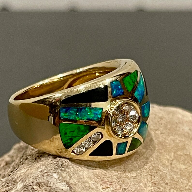 May include: A gold ring with a large, colorful stone set in the center. The stone is surrounded by smaller stones, and the ring is set with a black and green design.