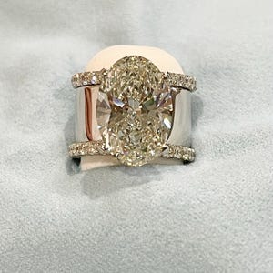 May include: A white gold engagement ring with a large oval-shaped diamond center stone and a diamond halo. The ring has a double band with pave diamonds.