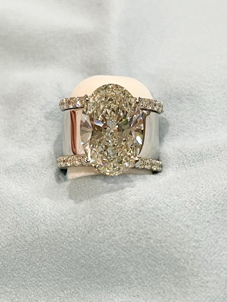 May include: A white gold engagement ring with a large oval-shaped diamond center stone and a diamond halo. The ring has a double band with pave diamonds.
