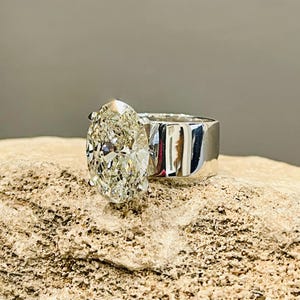 May include: A silver ring with a large, oval-shaped diamond set in a simple band. The ring is on a light brown, textured surface.