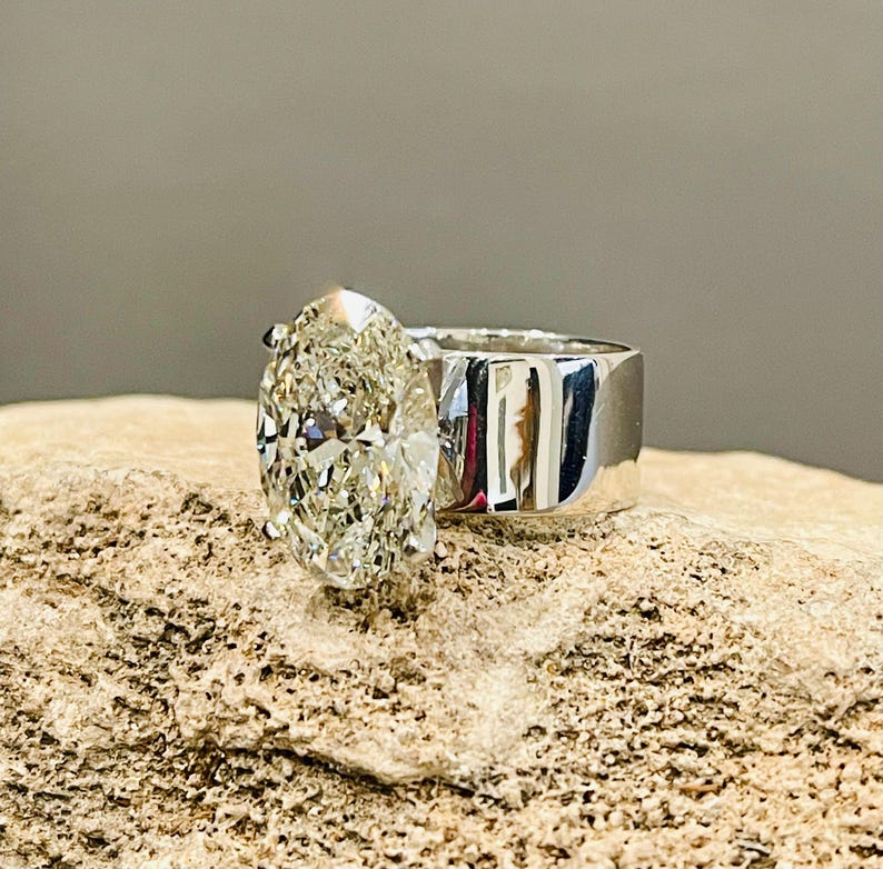 May include: A silver ring with a large, oval-shaped diamond set in a simple band. The ring is on a light brown, textured surface.