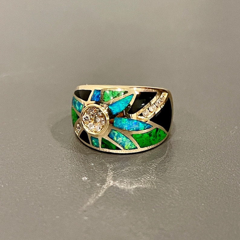 May include: A gold ring with a black, green, and blue inlay design. The ring features a round, clear stone in the center.