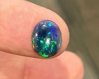 May include: A polished, oval-shaped black opal gemstone. The opal displays vibrant flashes of green, blue, and yellow colors within its dark body. The gemstone is held between two fingers, showcasing its iridescent play-of-color.