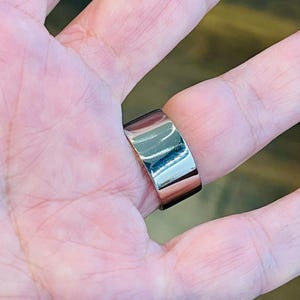 May include: A silver ring with a wide band. The ring is worn on a person's finger.