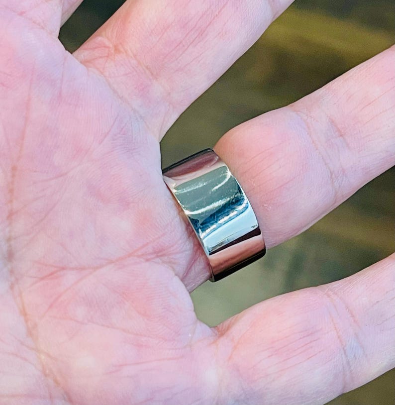 May include: A silver ring with a wide band. The ring is worn on a person's finger.