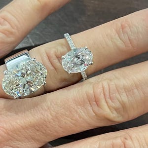 May include: Two oval-shaped diamond engagement rings on a hand. The larger ring has a thick band and a large center stone. The smaller ring has a thinner band and a smaller center stone.