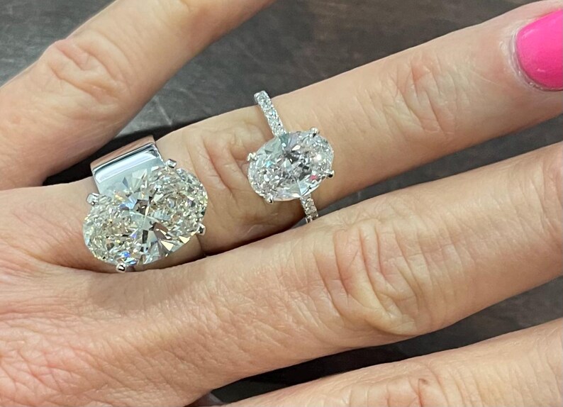 May include: Two oval-shaped diamond engagement rings on a hand. The larger ring has a thick band and a large center stone. The smaller ring has a thinner band and a smaller center stone.