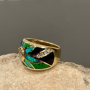 May include: A gold ring with a black and green opal inlay and a row of diamonds. The ring is set on a rock.