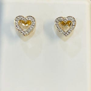 May include: A pair of heart-shaped stud earrings. Each earring features a gold-colored heart center surrounded by a halo of small, clear gemstones. The earrings are displayed against a white background.