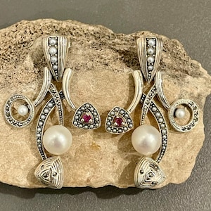 May include: A pair of silver earrings with a pearl and ruby accents. The earrings are made of silver and have a delicate design. The earrings are perfect for a special occasion.