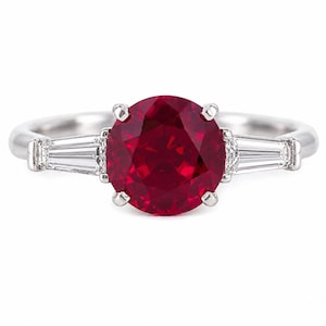 May include: A silver ring featuring a large, round, deep red gemstone set in the center with four prongs. On each side of the center stone, a tapered baguette-cut clear gemstone is set horizontally into the polished silver band.