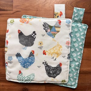 May include: Cream-colored pot holder with a patchwork chicken design. The chickens are in various colors and patterns, with a teal floral pattern on the back. A matching loop is attached for hanging.