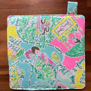 May include: A square, colorful pot holder with a vibrant print. The design features illustrations of women, fruit, and text, including "Juice Stand" and "1960". The color palette includes pink, blue, yellow, and green.