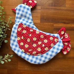 May include: A fabric chicken-shaped kitchen accessory featuring a light blue and white gingham pattern on the body. The wing is a red fabric patch with a white daisy and polka dot print. The comb and tail are made of the same red floral fabric.