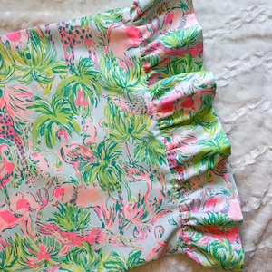 May include: A section of fabric with a tropical print featuring pink flamingos, giraffes, and palm trees on a light blue background. The fabric has a ruffled edge in pink and green.