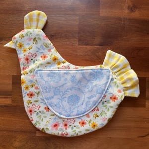 May include: A fabric chicken-shaped kitchen accessory featuring a cream-colored base with a multi-colored floral print. It includes a light blue patterned wing patch, a yellow and white gingham comb, and a matching yellow and white gingham tail.