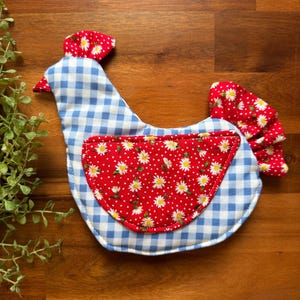 May include: A handmade chicken-shaped potholder. The body is blue and white gingham, with a red, white, and yellow floral pattern on the breast. The comb and tail are red with white polka dots. The potholder is on a wooden surface.