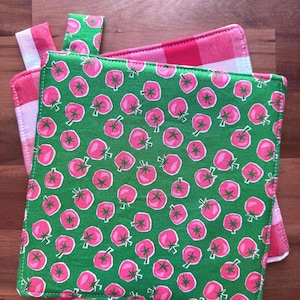 May include: A stack of kitchen textiles featuring a green tomato pattern. The main textile has a green background with pink tomato illustrations. A pink and white gingham pattern is visible on the edges and on a separate tab.