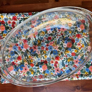 May include: A clear glass oval baking dish with integrated side handles, positioned over a quilted fabric mat featuring a dense, colorful floral pattern of red, pink, blue, and yellow flowers with green leaves on a white background.