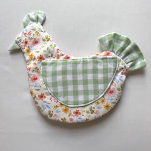 May include: A chicken-shaped fabric item featuring a white base with a colorful floral print of pink, orange, and yellow flowers. It includes a green and white gingham check fabric wing and tail, with a matching gingham comb on the head.