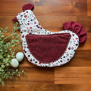 May include: A chicken-shaped oven mitt with a white floral pattern and a burgundy pocket and tail. The mitt is next to two light blue eggs and greenery on a wooden surface.