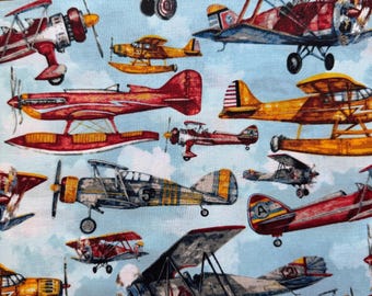 May include: A fabric pattern featuring various vintage airplanes in shades of red, yellow, and gray against a light blue sky with white clouds. The planes have propellers and wings, creating a retro aviation theme.