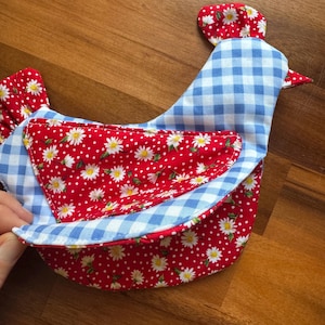 May include: A fabric chicken-shaped kitchen accessory featuring a blue and white gingham pattern on the body and a red fabric with a white daisy and polka dot print on the wing, tail, and comb. The item is constructed with layered textile panels.