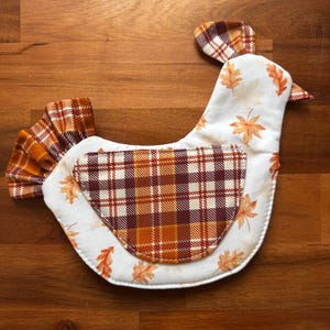 May include: A fabric chicken-shaped potholder featuring a white base with an orange and brown autumn leaf print. The wing and tail are made of a brown, orange, and white plaid fabric. The comb on the head also features the same plaid pattern.