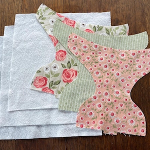 May include: A collection of fabric pieces for sewing projects. The image shows several white felt squares and patterned fabric cutouts in floral and geometric designs. The fabrics feature colors like pink, green, and cream.