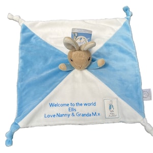 Puede incluir: Una manta para bebé azul y blanca con un peluche de Peter Rabbit adjunto. La manta tiene un mensaje personalizado que dice "Bienvenido al mundo Ellis Love Nanny & Granda M.x".