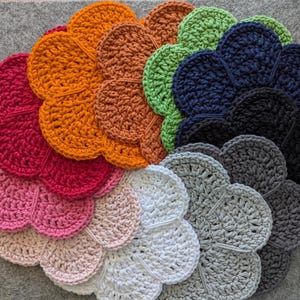 May include: A collection of handmade crochet flower-shaped coasters in various colors, including pink, red, orange, green, blue, gray, and black. The coasters are arranged in a circular pattern, showcasing the different hues and textures of the yarn.