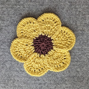 May include: A crocheted sunflower coaster in yellow and brown. The coaster is made of yarn and has a flower shape with a brown center.