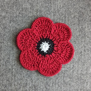 May include: A red crocheted poppy flower with a black and white center. The flower is made with a single crochet stitch and has a textured appearance.
