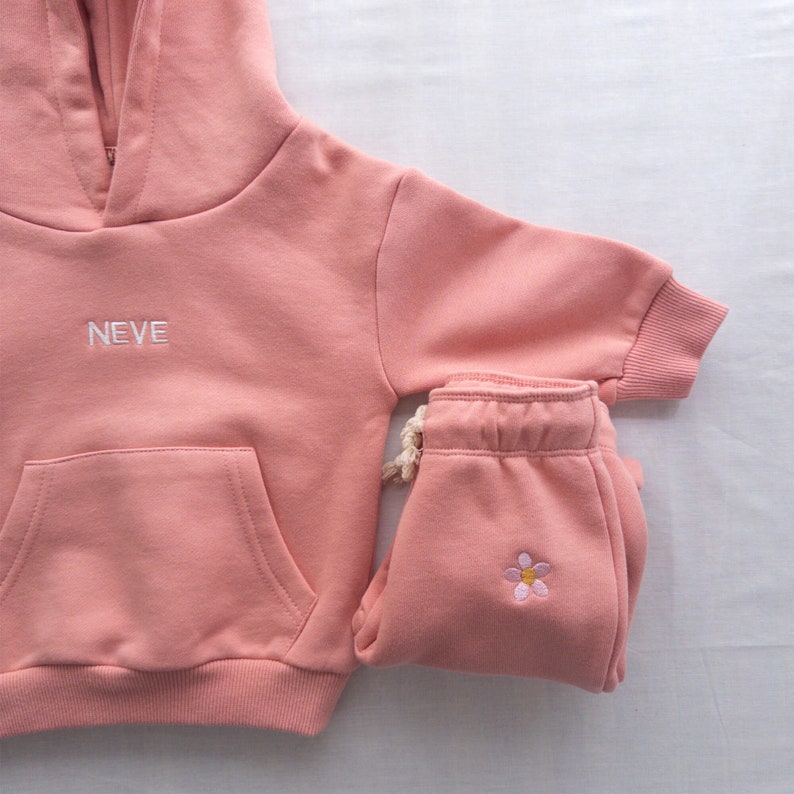 May include: A pink hooded sweatshirt with the word "NEVE" embroidered on the front and a matching pair of pink sweatpants with a small embroidered flower on the left leg.