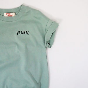 以下が含まれることがあります： 薄い緑色のTシャツで、前面に黒い刺繍で「JOANIE」と書いてあります。Tシャツには、「Tiny Kind」と書かれた小さな赤いタグが付いています。