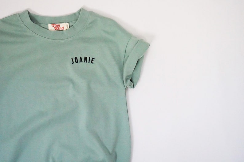 以下が含まれることがあります： 薄い緑色のTシャツで、前面に黒い刺繍で「JOANIE」と書いてあります。Tシャツには、「Tiny Kind」と書かれた小さな赤いタグが付いています。