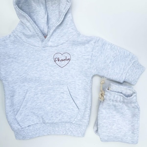 May include: A gray hooded sweatshirt with a heart-shaped design that says "Phoebe" in a burgundy color. The sweatshirt is paired with matching gray sweatpants.