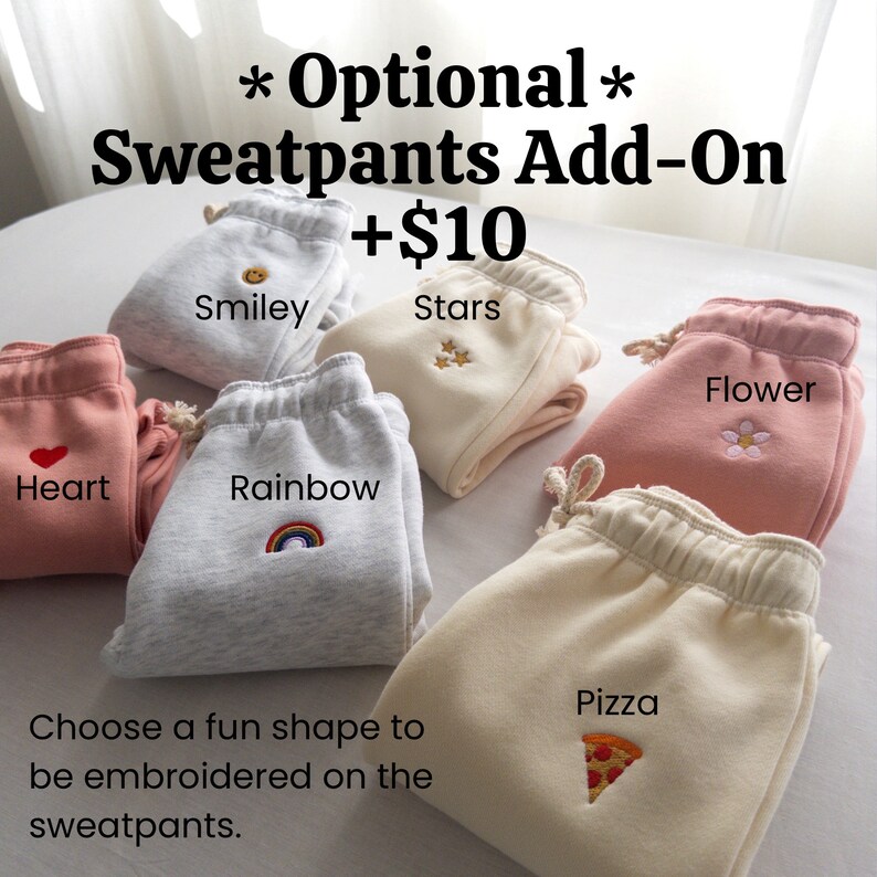 May include: Six pairs of sweatpants with different embroidered designs: a smiley face, stars, a heart, a rainbow, a flower, and a pizza slice. The text "Optional Sweatpants Add-On +$10" is at the top of the image. The text "Choose a fun shape to be embroidered on the sweatpants." is at the bottom of the image.