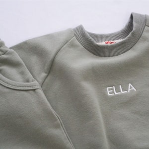 Pode incluir: Uma camisola de cor verde-s&aacute;lvia com o nome "ELLA" bordado a branco na frente, combinada com cal&ccedil;as de fato de treino a condizer de cor verde-s&aacute;lvia.