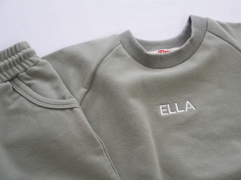 Pode incluir: Uma camisola de cor verde-s&aacute;lvia com o nome "ELLA" bordado a branco na frente, combinada com cal&ccedil;as de fato de treino a condizer de cor verde-s&aacute;lvia.