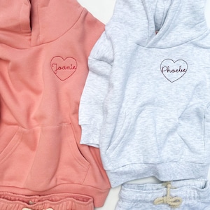 May include: Two hooded sweatshirts with heart-shaped embroidery. One is pink with the name "Joanie" embroidered in red, the other is gray with the name "Phoebe" embroidered in red.
