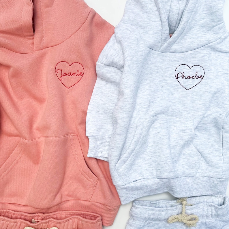 May include: Two hooded sweatshirts with heart-shaped embroidery. One is pink with the name "Joanie" embroidered in red, the other is gray with the name "Phoebe" embroidered in red.