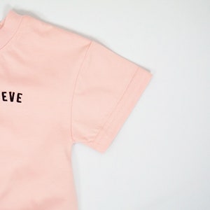以下が含まれることがあります： 前面に黒で「NEVE」とプリントされたピンクの半袖Tシャツです。