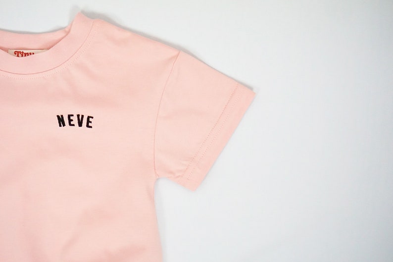 以下が含まれることがあります： 前面に黒で「NEVE」とプリントされたピンクの半袖Tシャツです。