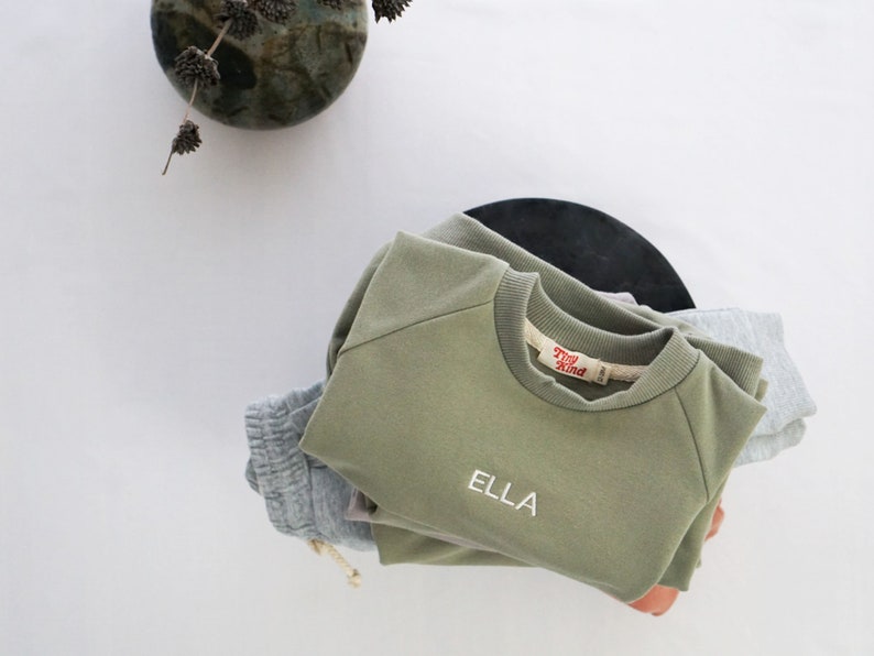 Pode incluir: Uma pilha de roupas infantis em verde e cinza com o nome "Ella" bordado na camiseta verde. A camiseta tem uma etiqueta vermelha que diz "Tiny Kind".